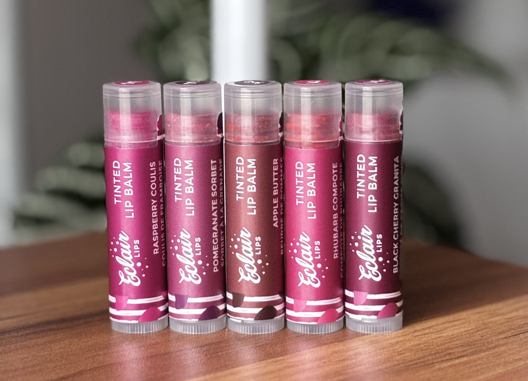 Five Eclair Lips tinted lip balms, featuring Raspberry Coulis, Pomegranate Sorbet, Apple Butter, Rhubarb Compote, and Black Cherry Granita flavors, are neatly arranged on a wooden table.