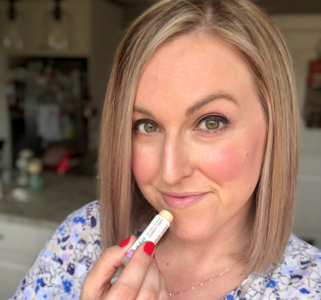A smiling woman with blonde hair and green eyes holds an Eclair Lips Candy Tart natural lip balm stick, showing the product clearly with red nail polish, against a blurred indoor background.