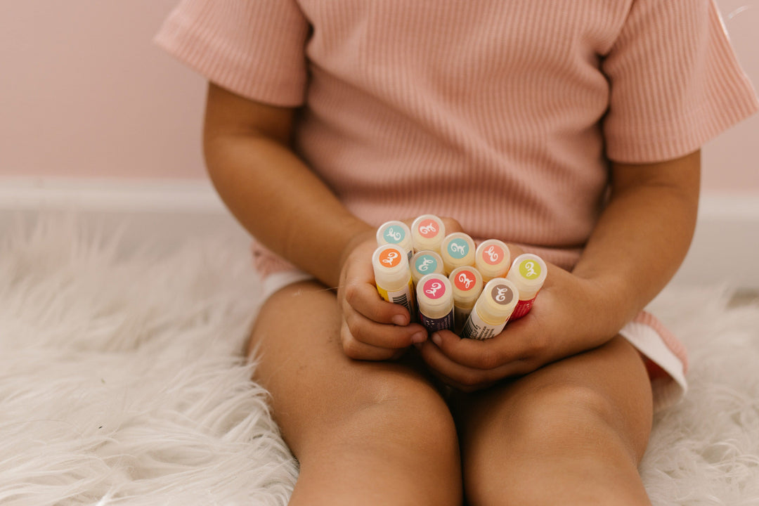 A child with light brown skin holds a cluster of colorful Eclair Lips lip balm tubes while sitting on a fluffy white rug.