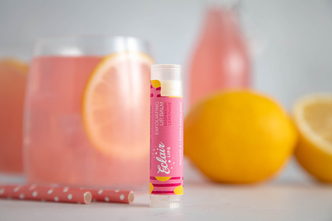 A vibrant pink Eclair Lips exfoliating lip balm in lemonade flavor stands upright on a white surface, with a glass of pink lemonade and fresh lemons blurred in the background.