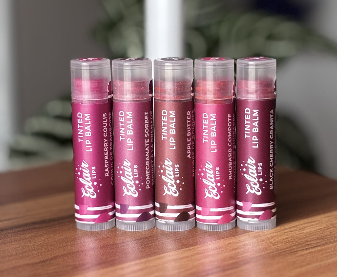 Five Eclair Lips tinted lip balms, featuring Raspberry Coulis, Pomegranate Sorbet, Apple Butter, Rhubarb Compote, and Black Cherry Granita flavors, are neatly arranged on a wooden table.