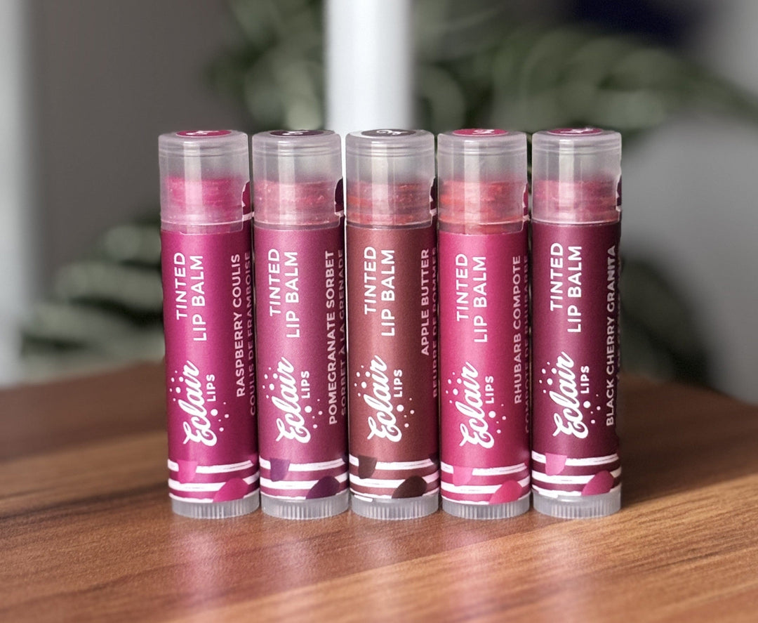 Five Eclair Lips tinted lip balms, featuring Raspberry Coulis, Pomegranate Sorbet, Apple Butter, Rhubarb Compote, and Black Cherry Granita flavors, are neatly arranged on a wooden table.