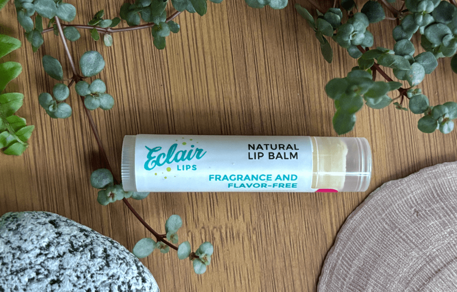 A white Eclair Lips Natural Lip Balm, labeled 'Fragrance and Flavor-Free,' is displayed on a wooden surface with green plants and a textured grey rock.