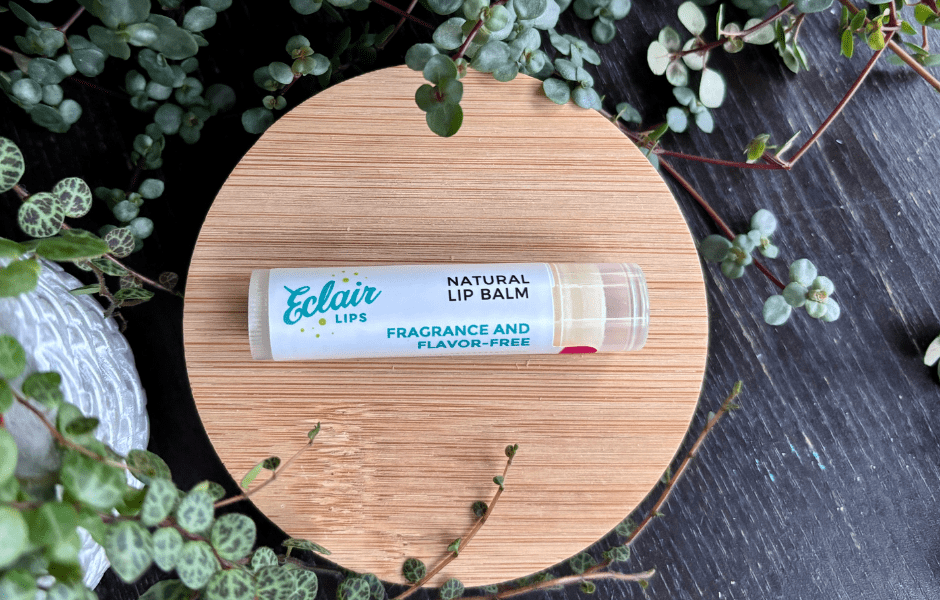 A white Eclair Lips Natural Lip Balm, labeled 'Fragrance and Flavor-Free,' is displayed on a wooden surface with green plants and a textured grey rock.