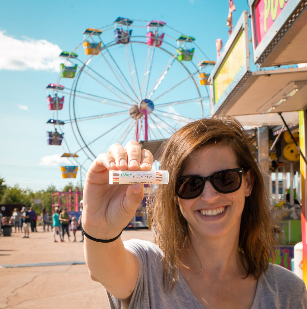 A smiling woman in sunglasses holds up an Eclair Lips Funnel Cake lip balm in front of a blurry carnival Ferris wheel and food stalls on a sunny day.