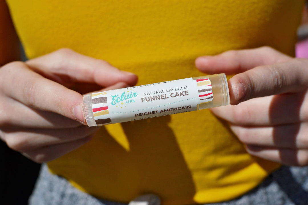 A tube of Eclair Lips Funnel Cake lip balm held with the index fingers of both hands in front of a person's torso clad with a yellow top