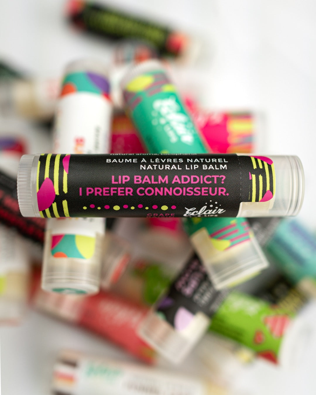 A clear tube of Eclair Lips natural grape lip balm with a black label featuring colorful dots rests horizontally across a textured black and plain yellow background.