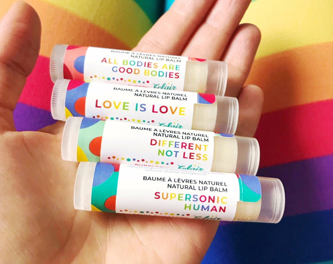 A person's hand holds four Éclair natural lip balms with empowering messages like 'LOVE IS LOVE' and 'ALL BODIES ARE GOOD BODIES' against a vibrant rainbow background.