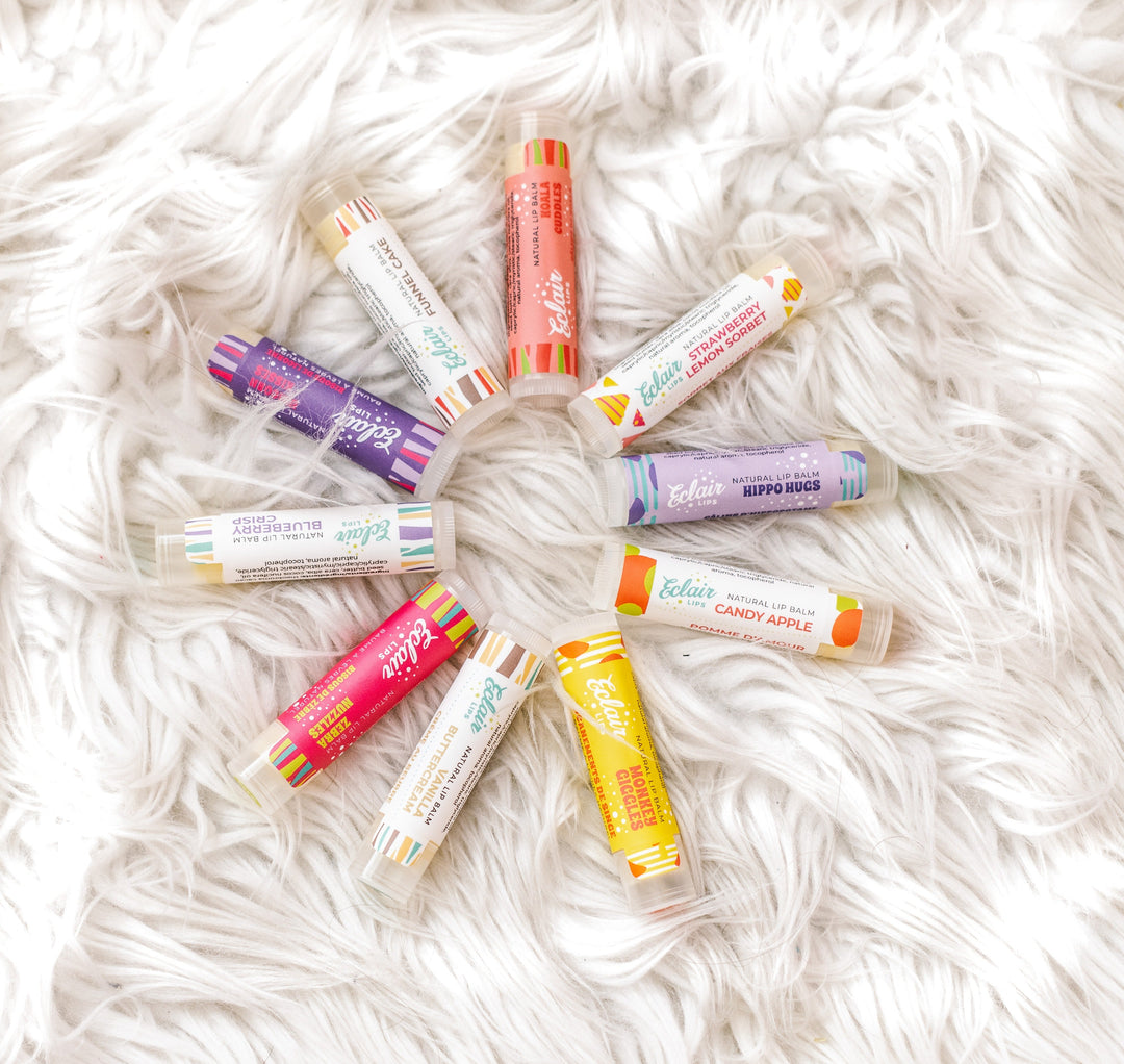 A flat lay product shot featuring ten Eclair Lips natural lip balms arranged in a circular pattern on a fluffy white faux fur surface. Each lip balm has a unique colorful label with playful names.