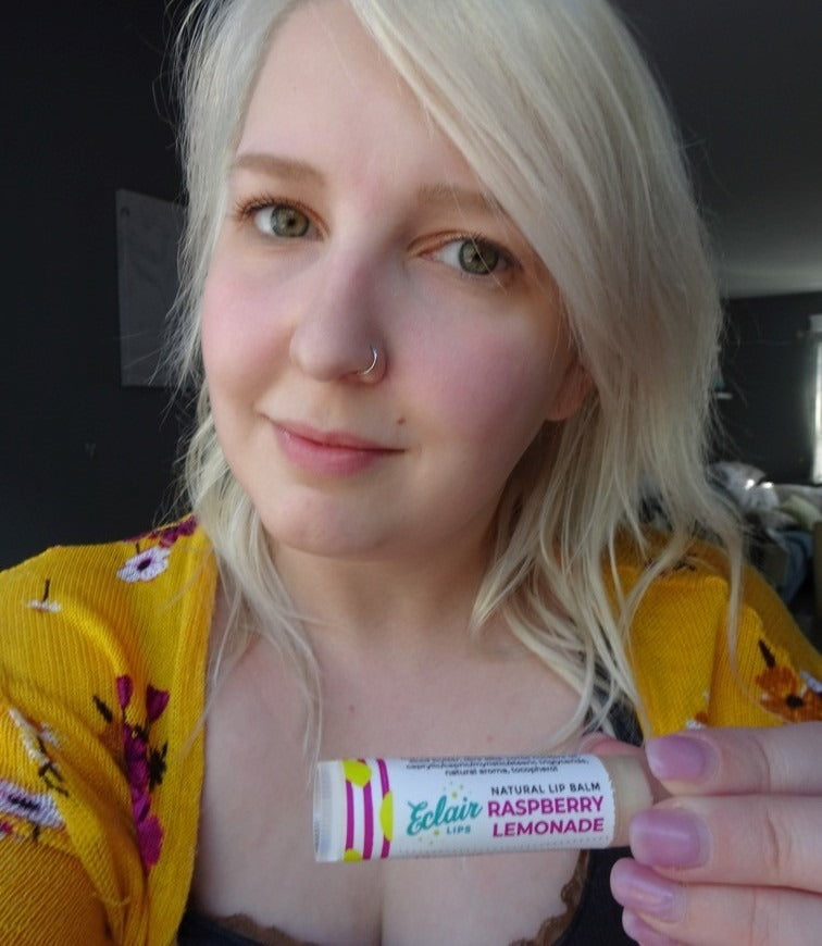 A young woman with blonde hair and a nose piercing smiles while holding up a tube of eclair LIPS Natural Lip Balm in Raspberry Lemonade flavor.