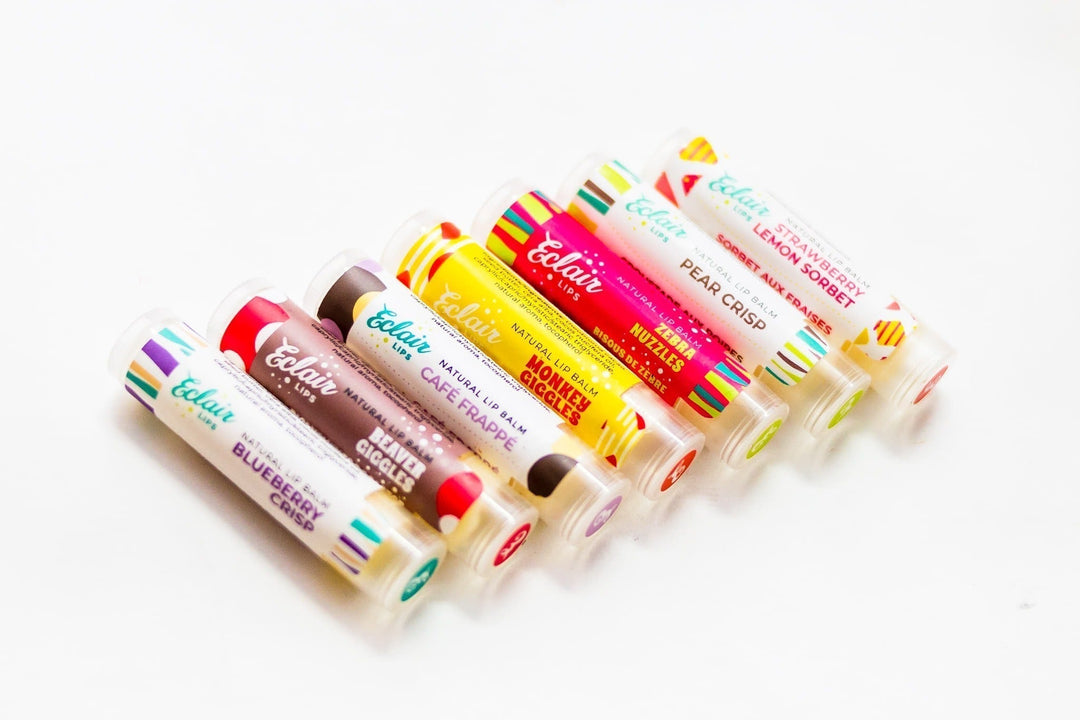 Seven colorful Éclair Lips brand lip balm tubes, in flavors such as Blueberry Crisp, Monkey Smiles, Cafe Frappe, and Lemon Sunset, arranged diagonally on a white background.