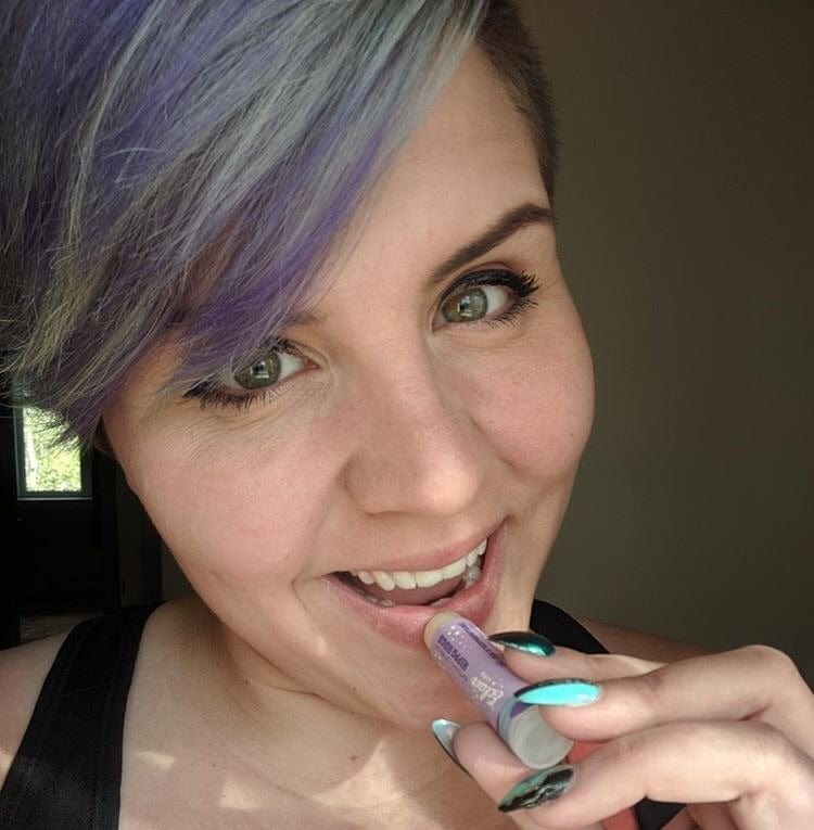 A smiling woman with purple and grey hair applies eclair lips lip balm, showcasing her bright green eyes and artistic turquoise and black nail polish.