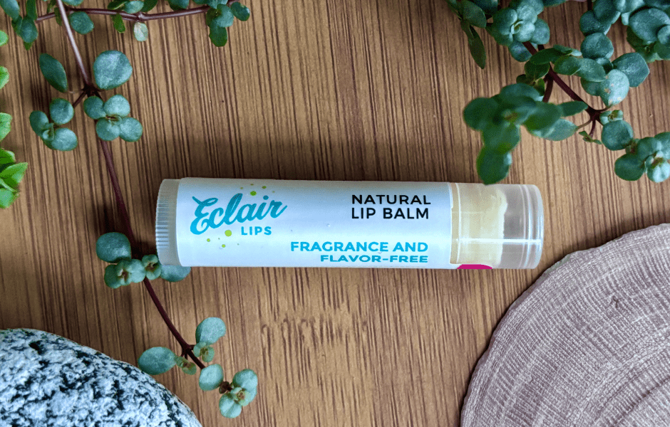 A white tube of Eclair LIPS Natural Lip Balm, labeled 'Fragrance and Flavor-Free,' lies on a wooden surface next to green plants, a grey stone, and a light pink shell.