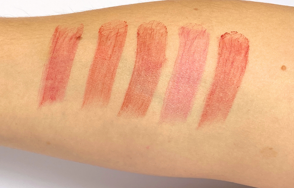 Six swatches of various pink and red lipsticks or tinted lip balms applied in vertical stripes on a person's fair-skinned arm.