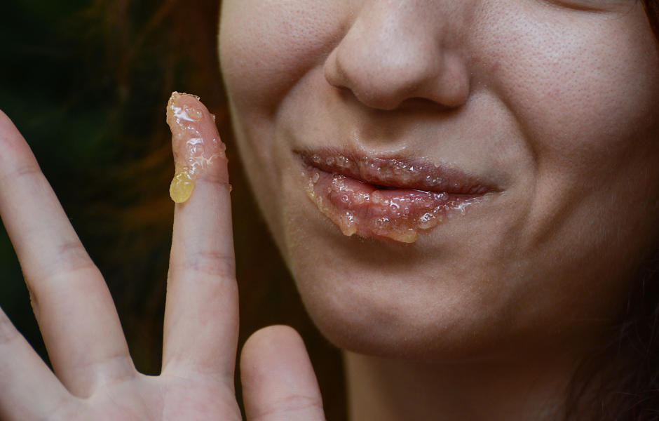 Close-up of a person's finger applying a glistening, grainy lip scrub to their lips, which are also covered in the same substance.