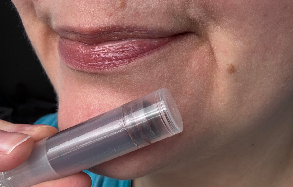 A close-up shot of a person with moisturized lips holding a clear tube of tinted lip balm near their mouth.