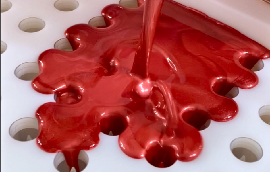 A vibrant, glossy tinted lip balm liquid is being poured from above onto into a lip balm filling tray spreading to fill the individual lip balm tubes underneath in a production process.