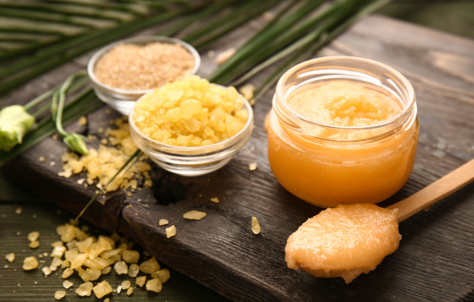 A jar of orange-colored body scrub, a wooden spoon with a dollop of scrub, and two small bowls containing yellow bath salts and brown sugar, all neatly arranged on a dark wooden surface